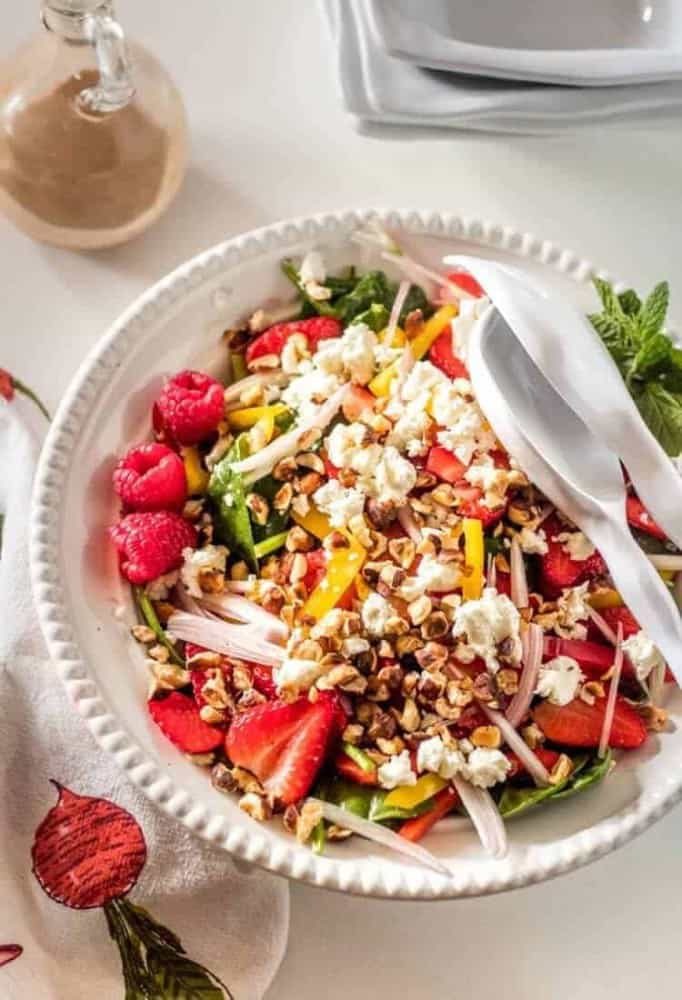 A spinach, strawberry, hazelnut, and chevre salad with Raspberry Fresh Mint Vinaigrette in a white ceramic bowl with white salad tongs.