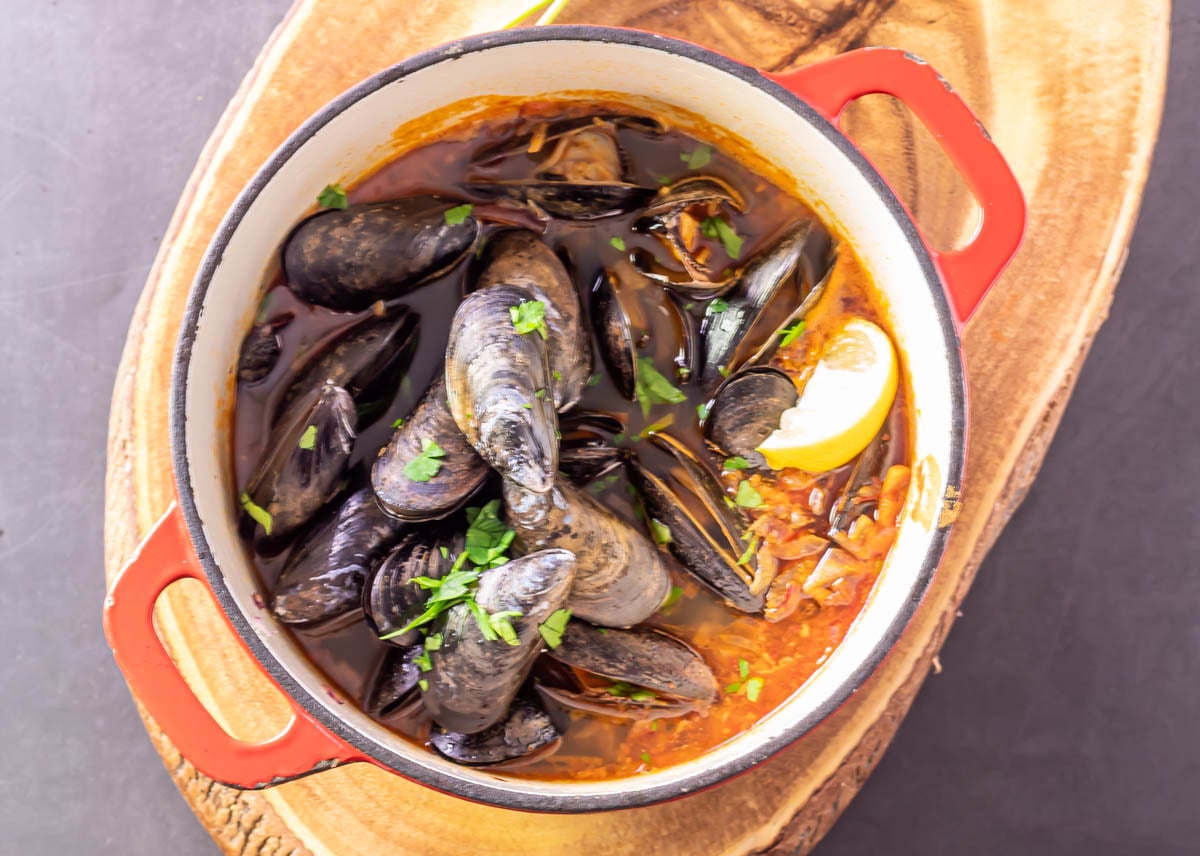 A red cast iron pot with the finished mussels garnished with parsley and a lemon wedge.