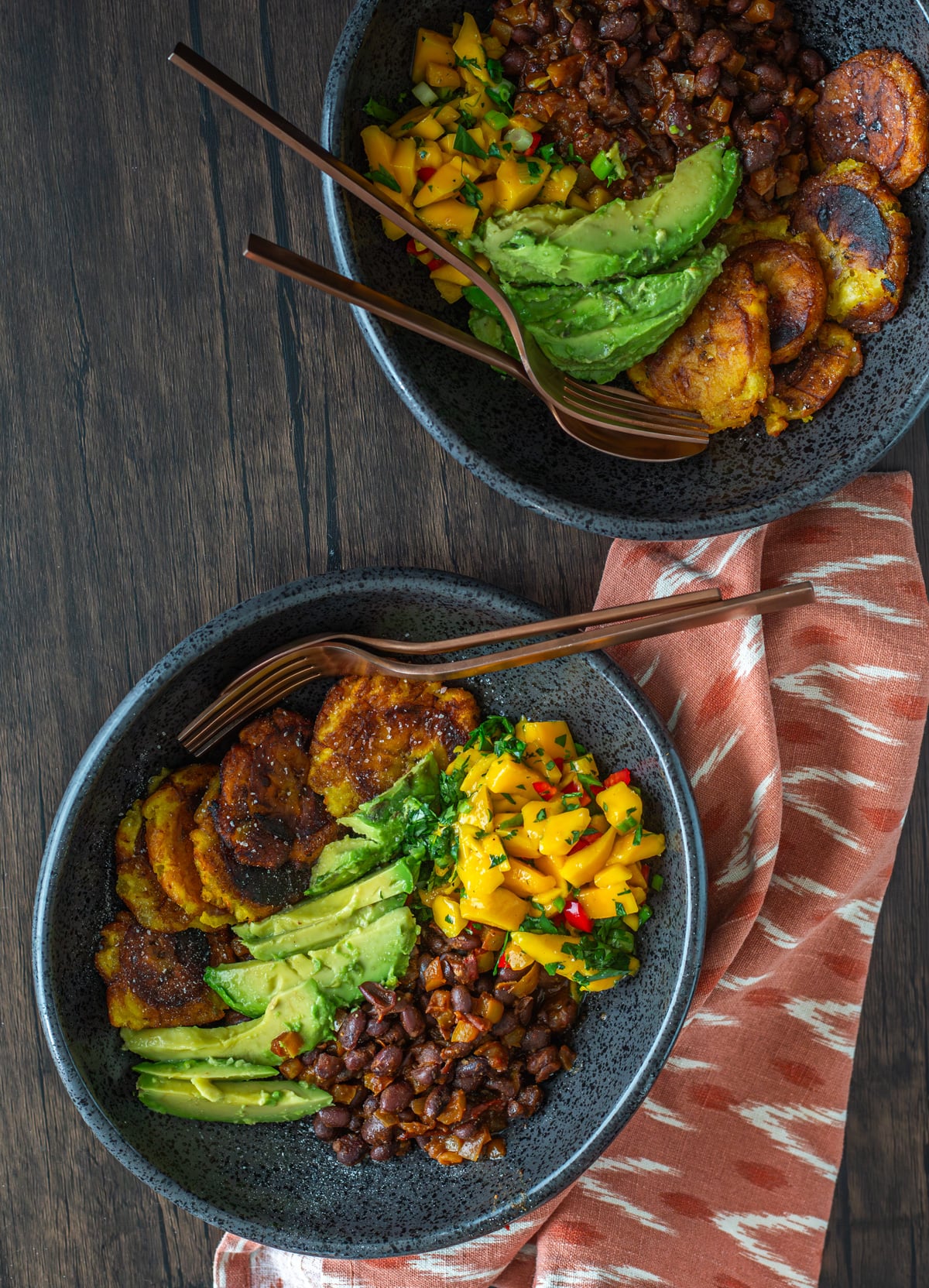 Tropical Buddha Bowl with Plantains, Black Beans, and Mango Salsa in black bowls with limes and cilantro on a brown background.