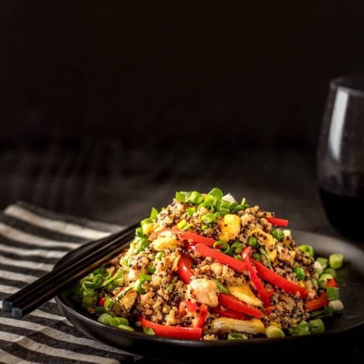 Quinoa Chaufa with chicken on a black plate with black chopsticks, a striped napkin, and a glass of wine