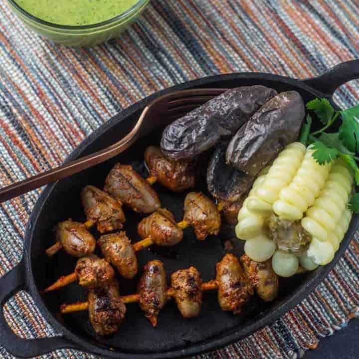 An oval cast iron plate with 3 Peruvian chicken anticuchos alongside roasted purple potatoes, choclo (giant corn), and 2 sauces - aji verde and rocoto aioli.