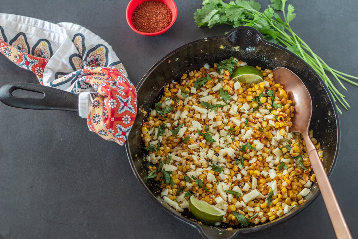 Taj&iacute;n corn in a skillet with copper spoon, fresh cilantro, and taj&iacute;n.