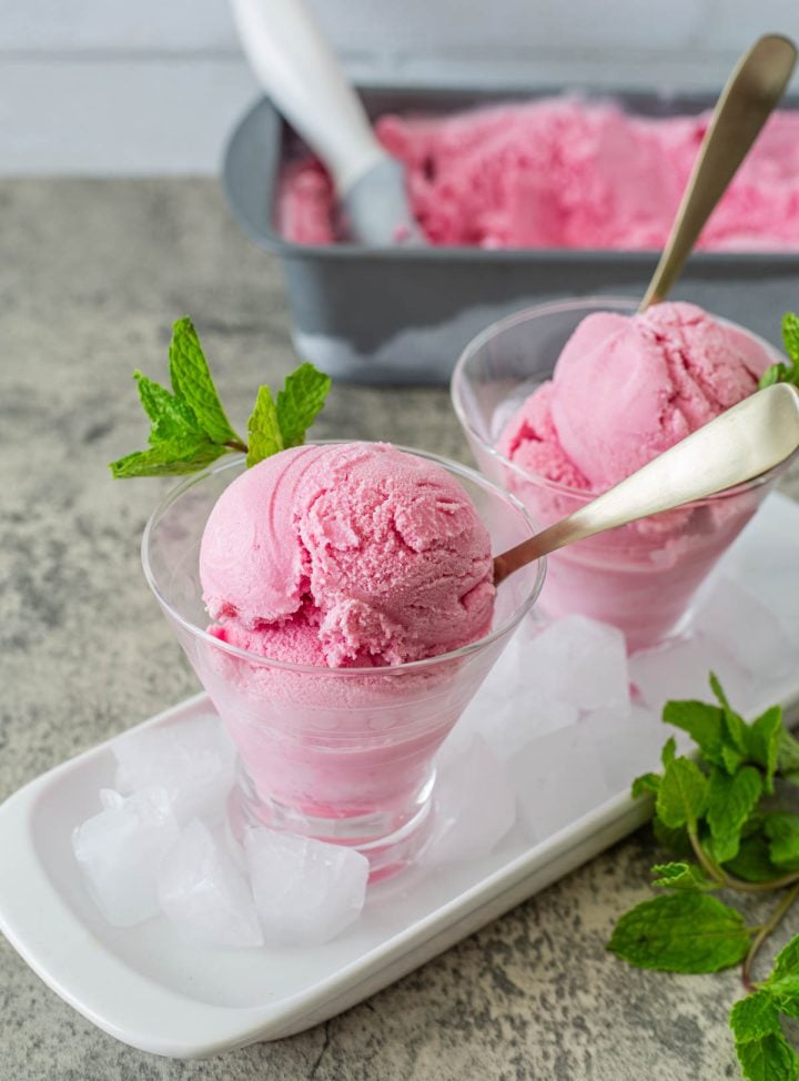 Prickly Pear Ice Cream in glassware on a tray with ice, and pan with custard ice cream and scoop in the background.