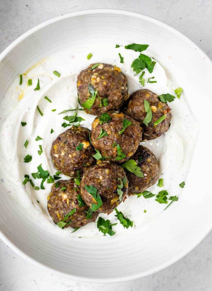 A white platter with Greek-style ground lamb meatballs on top of yogurt with chopped fresh herbs.