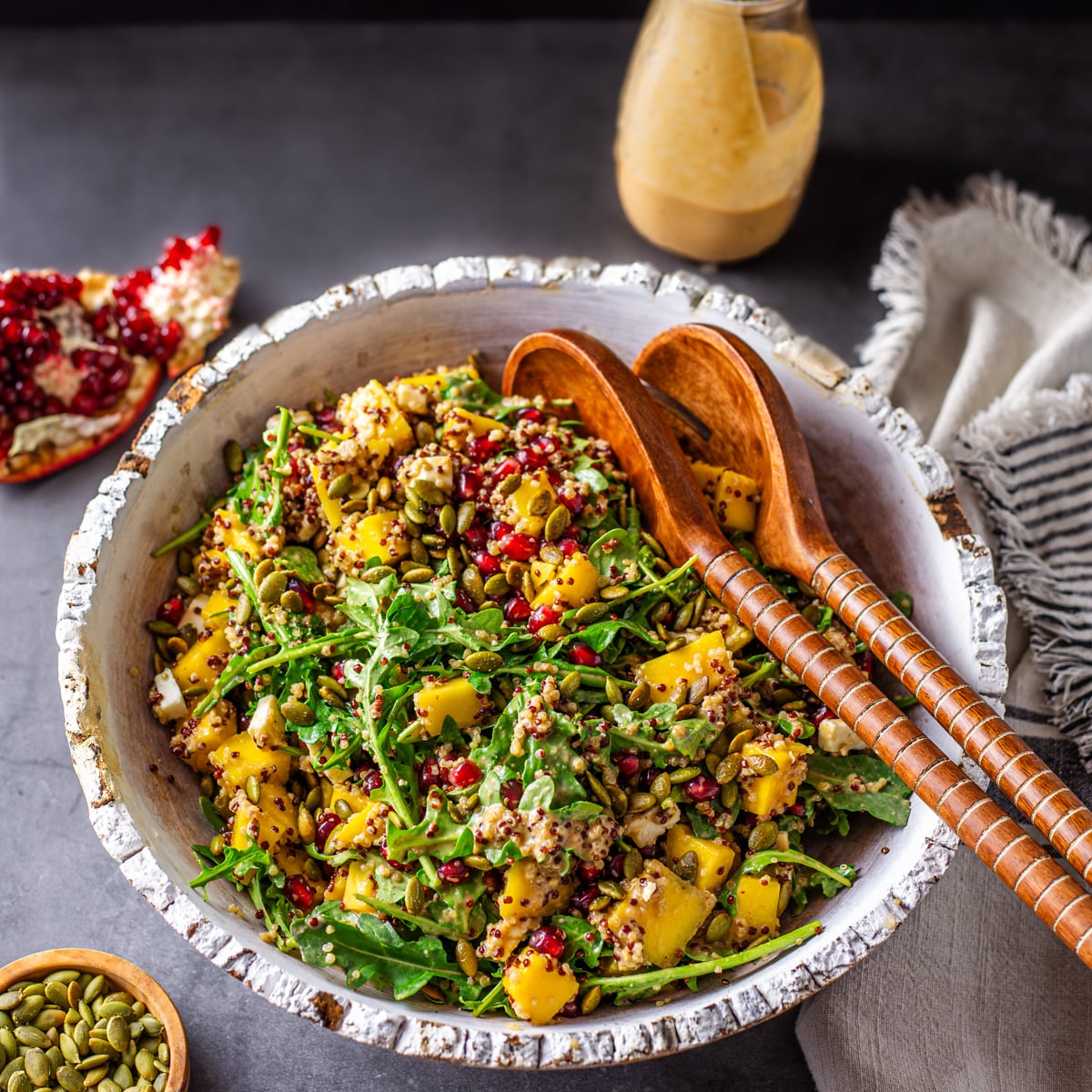 A rustic white wood bowl with quinoa and arugula salad with dressing and pepitas.