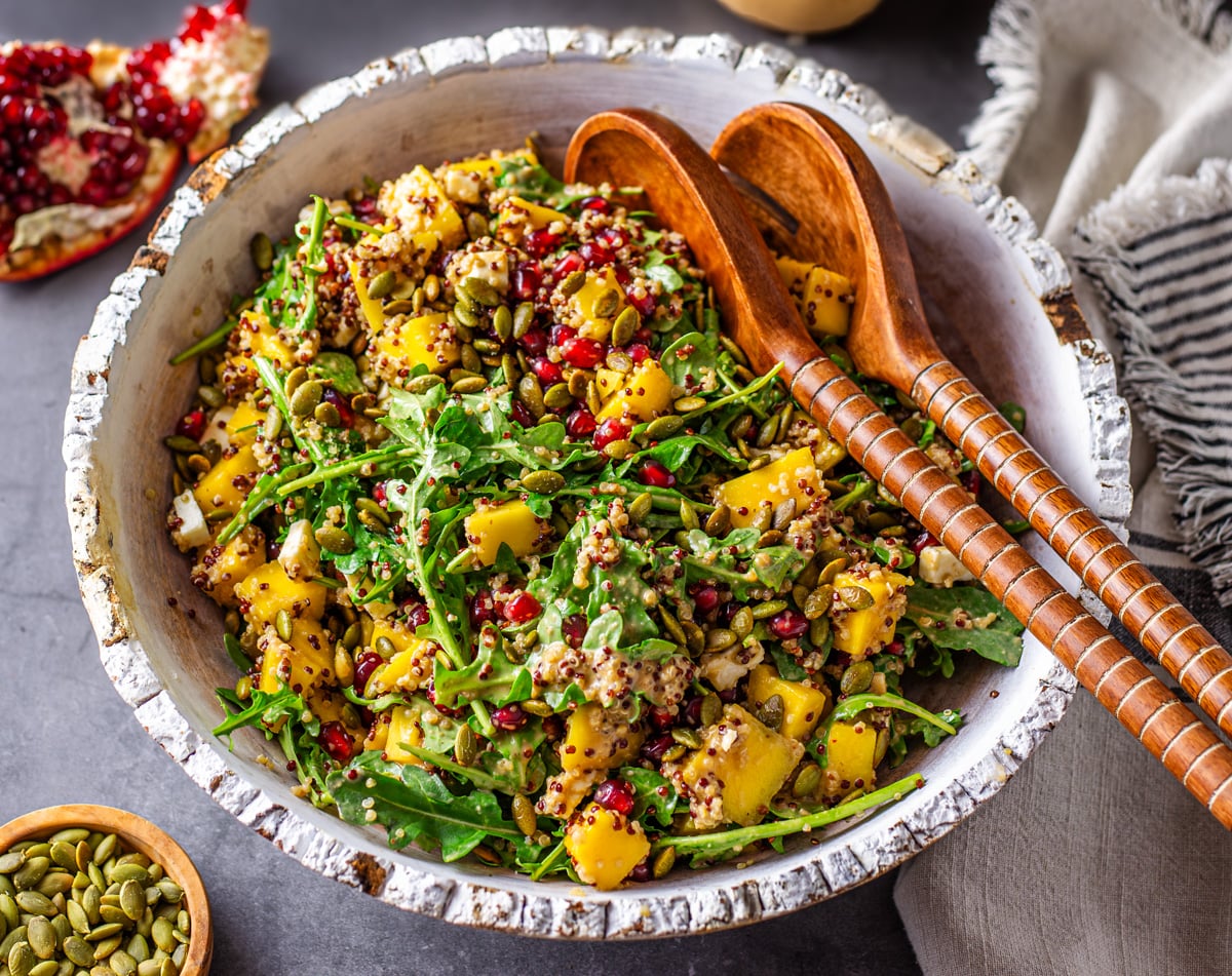 A close up of the quinoa and arugula salad with wood salad utensils, small bowl of pepitas, and part of a pomegranate.