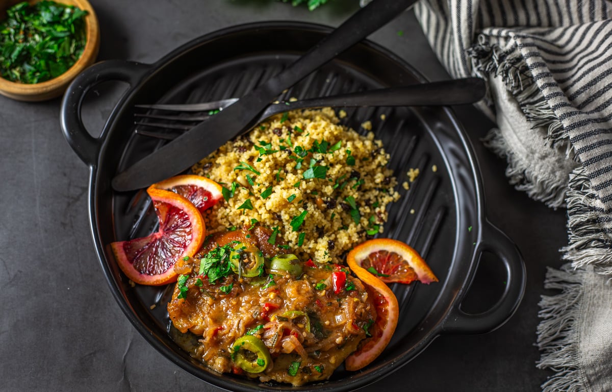 A black ceramic plate with Mediterranean blood orange chicken and couscous with orange slices.