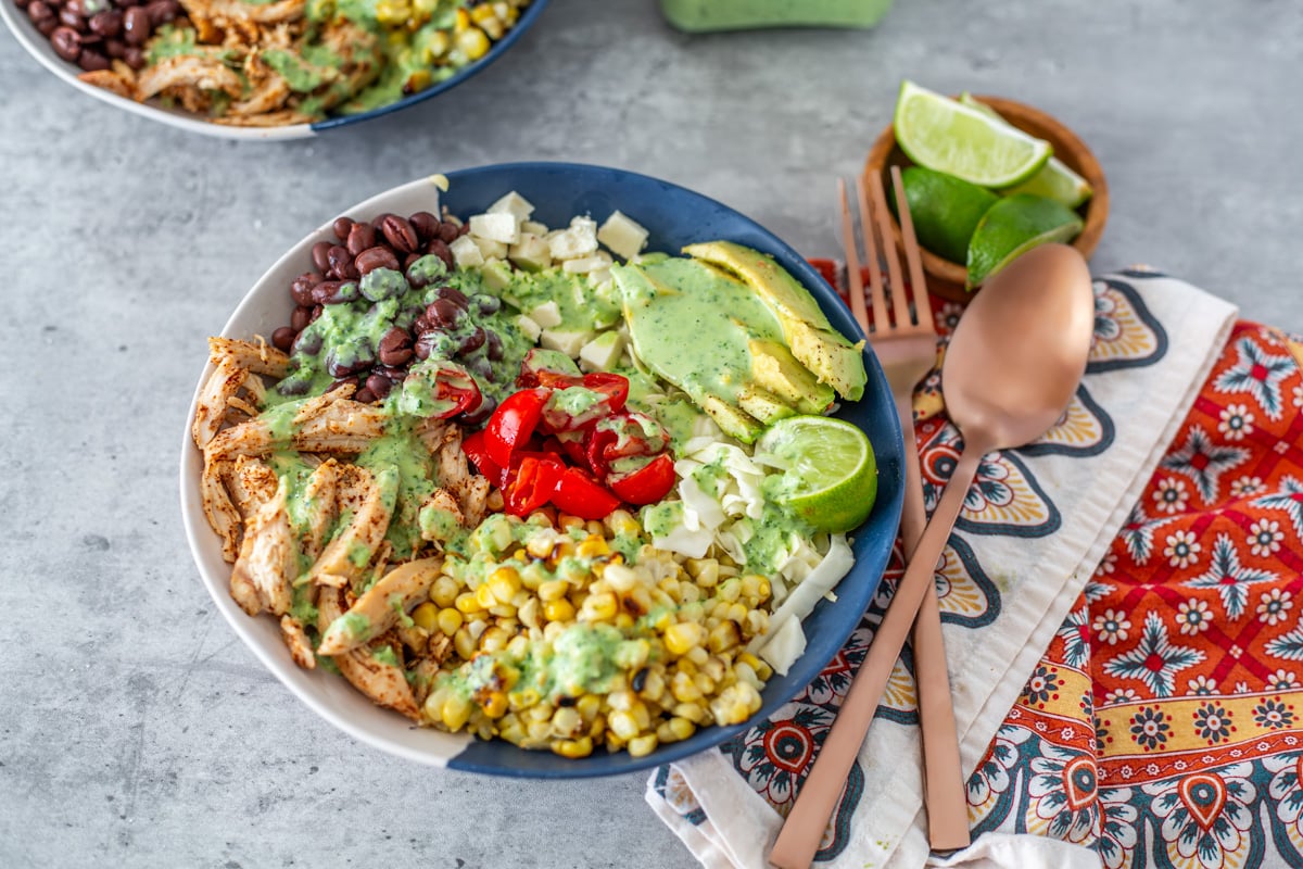 A close up of one blue and white bowl with taco-inspired ingredients, cloth napkin, and copper flatware.