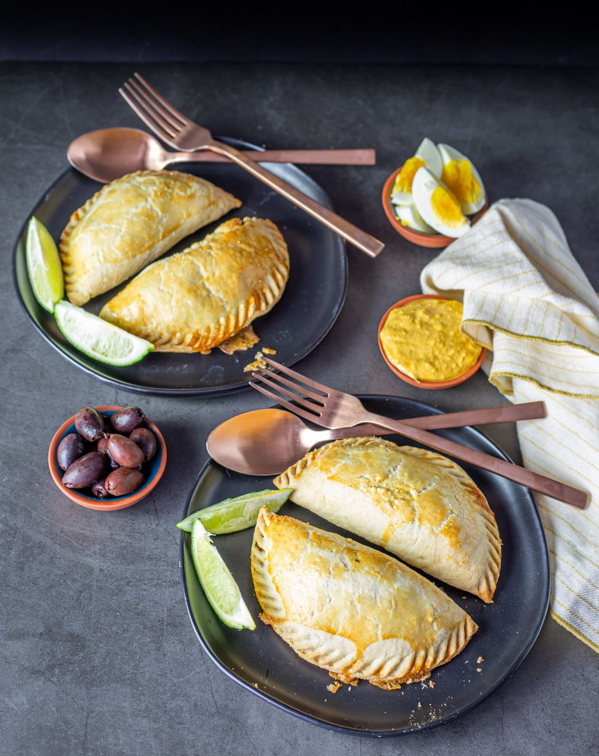 Two black ceramic plates with Peruvian beef empanadas, copper flatware, and garnishes.
