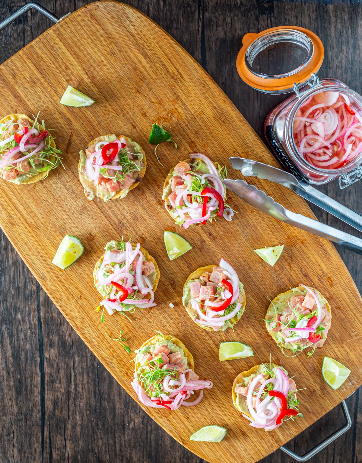 A wood cutting board with 8 appetizer-sized tostadas with tongs and pickled onion.