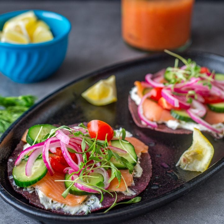 A black plate with 2 smoked salmon tostadas, lemon wedges, and a bloody mary.