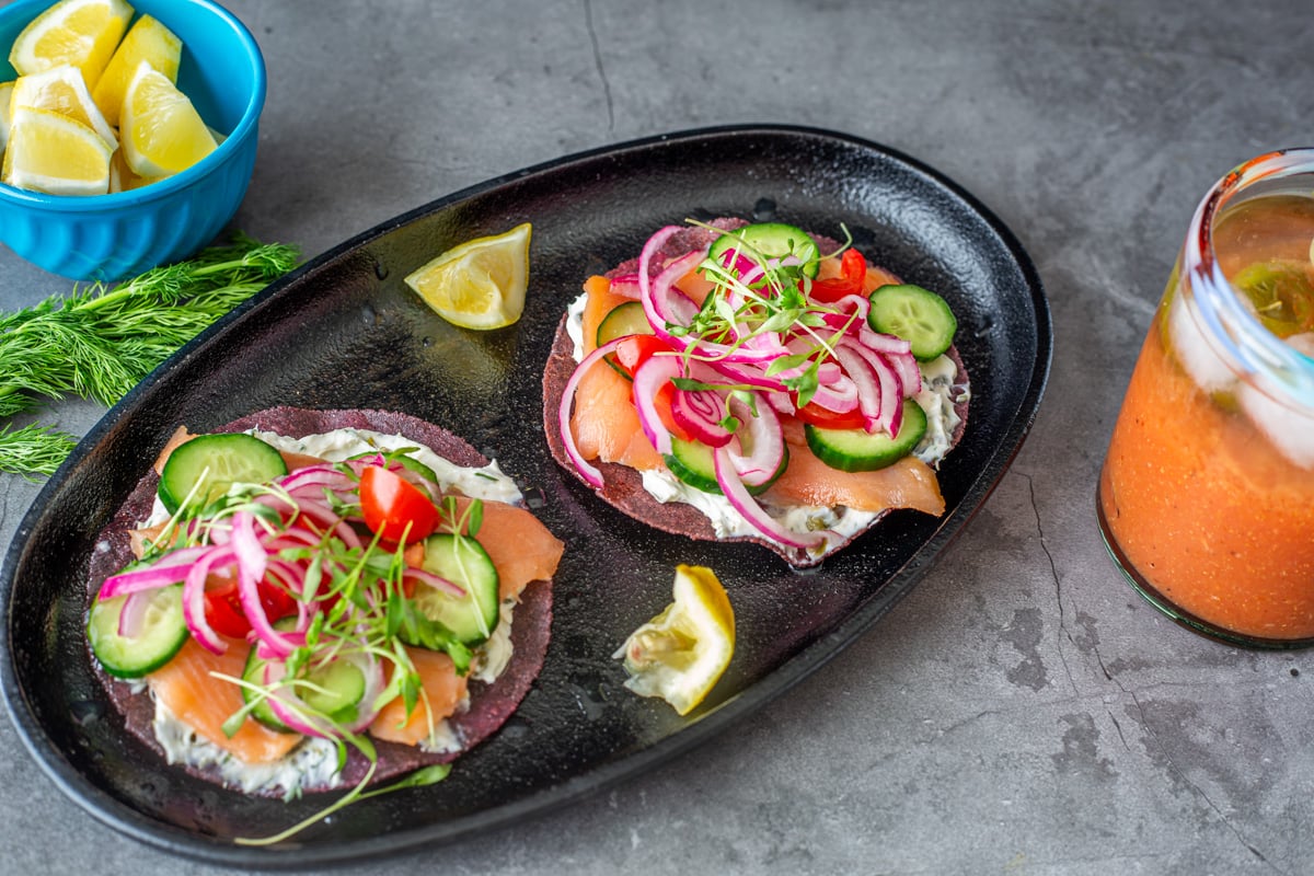 A close up of two tostadas on a black oval plate with a bloody mary and lemon wedges.