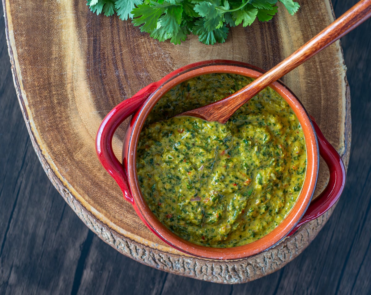 A red ceramic bowl with chimichurri sauce with cilantro and parsley, and wood spoon on a live edge wood cutting board.