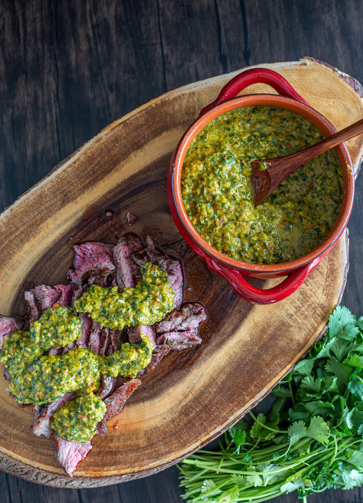 A wood cutting board with steak topped with cilantro chimichurri sauce, and a red ceramic bowl with extra sauce.