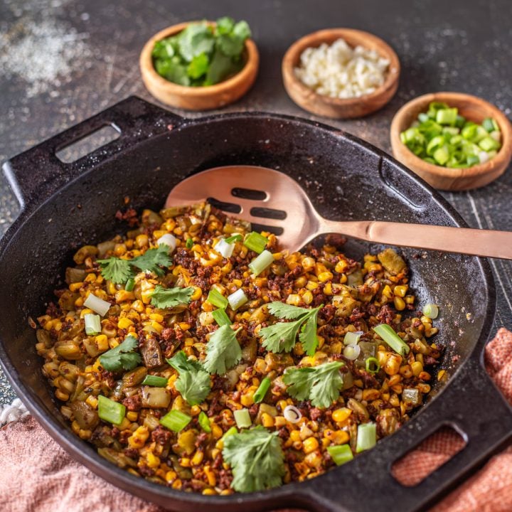A cast iron skillet with nopalitos with chorizo and corn with garnishes and copper spoon.