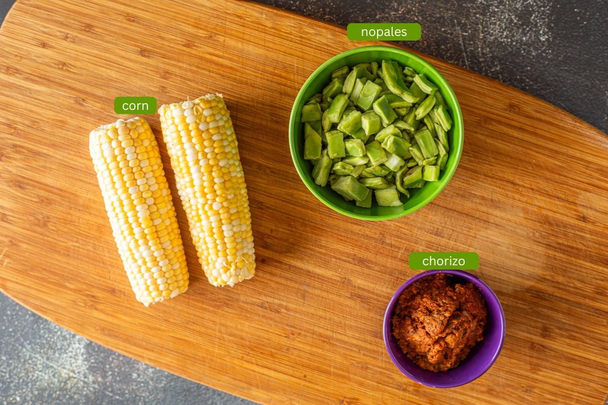 Ingredients for nopalitos with chorizo including fresh corn, nopales, and chorizo, on a wood cutting board.