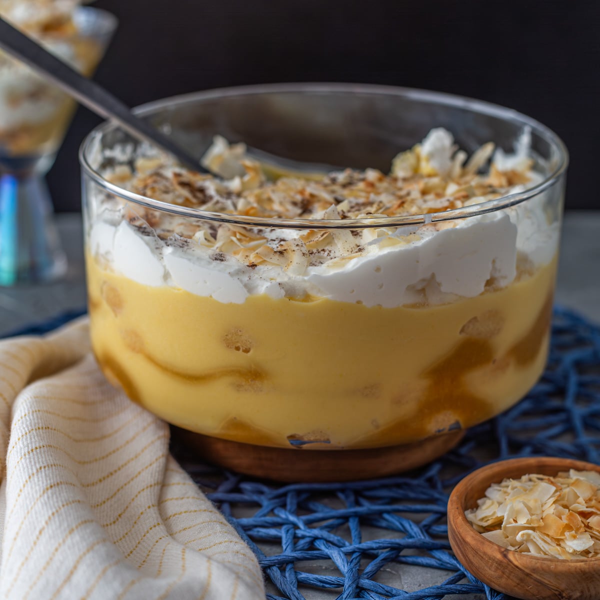 A glass trifle bowl with passionfruit trifle, cloth napkin, and small wood bowl of toasted coconut.