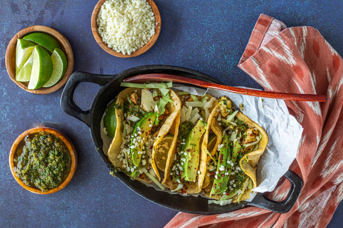 A close up of 3 chimichurri tacos in a cast iron plate with toppings and cloth napkin.