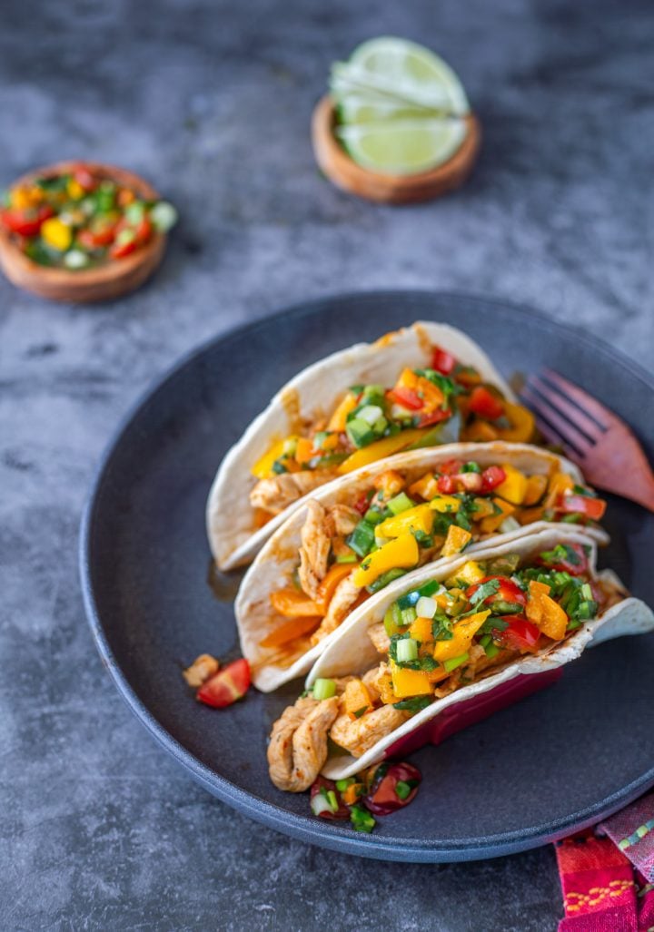 A charcoal grey ceramic plate with 3 Thai chicken curry tacos, mango salsa, and lime wedges.