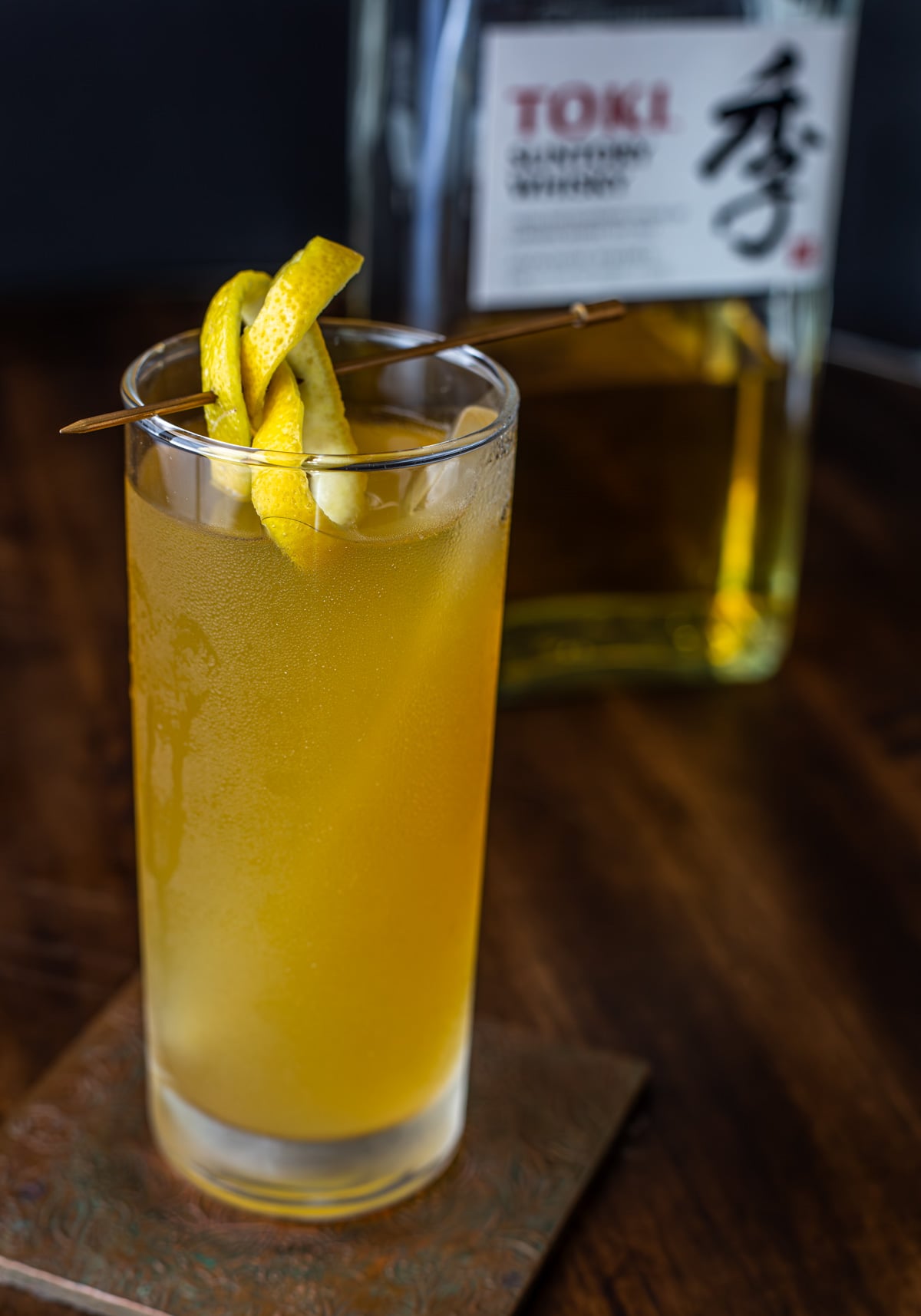 A highball glass with a whisky and green tea cocktail garnished with lemon peel.