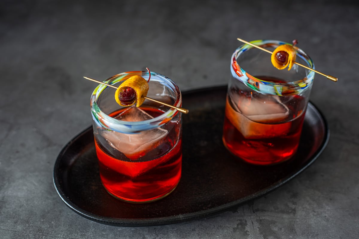 An oval cast iron tray with 2 mezcal negronis in Mexican rocks glasses with orange and cherry garnish.