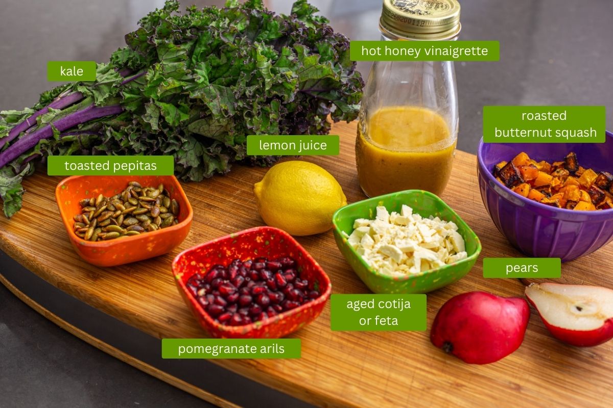 Ingredients for fall kale salad including fresh purple kale, hot honey vinaigrette, roasted squash, pears, cotija, pomegranate arils, pepitas.