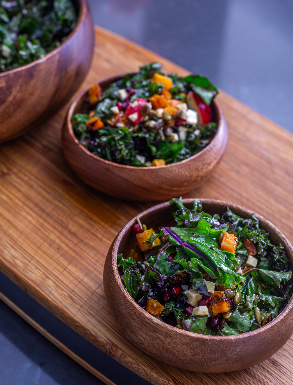 2 wooden salad bowls with fall kale salad and a large bowl of the salad in the background.