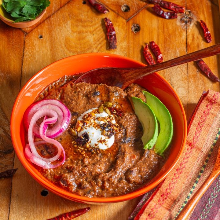An orange ceramic bowl with black bean soup, sour cream, salsa macha, avocado, and pickled onions.