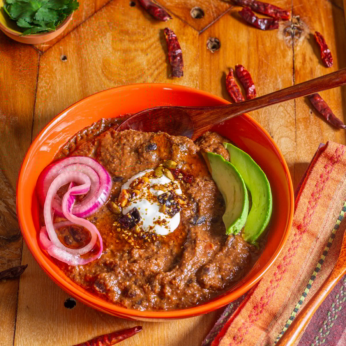 An orange ceramic bowl with black bean soup, sour cream, salsa macha, avocado, and pickled onions.