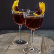 A close up of two prepared black manhattans with orange peel garnish on a wood board.