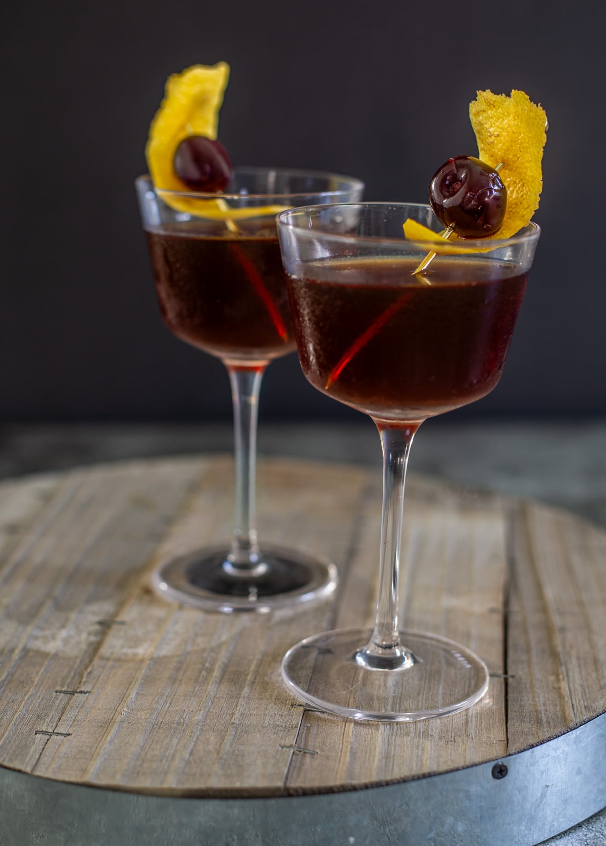 A wood board with 2 nocino cocktails garnished with orange peel and cherry.