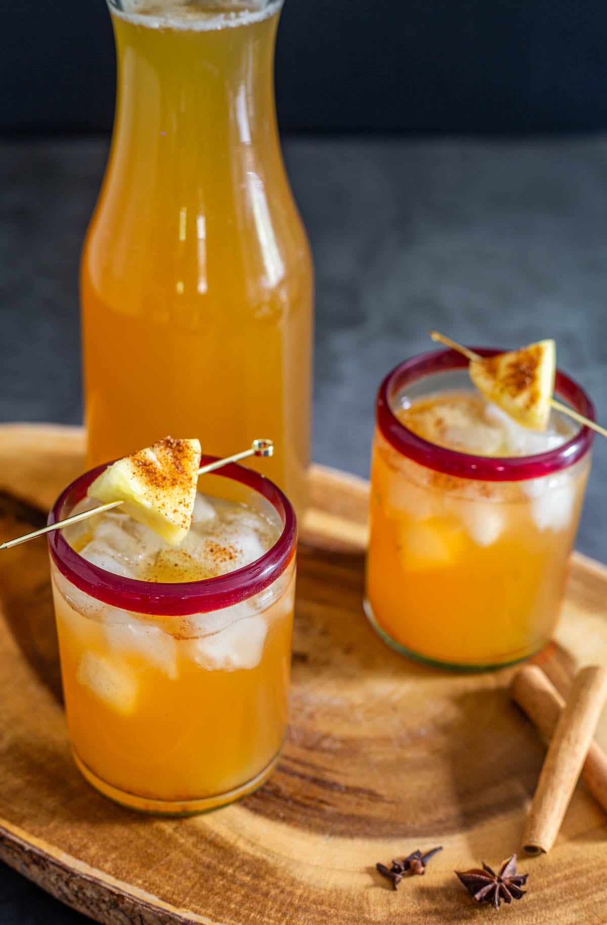 A carafe of homemade tepache with 2 red-rimmed glasses with ice, tepache, and pineapple cube.