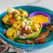 A turquoise ceramic bowl with 2 palta rellena halves, salsa huanca&iacute;na, and copper fork.