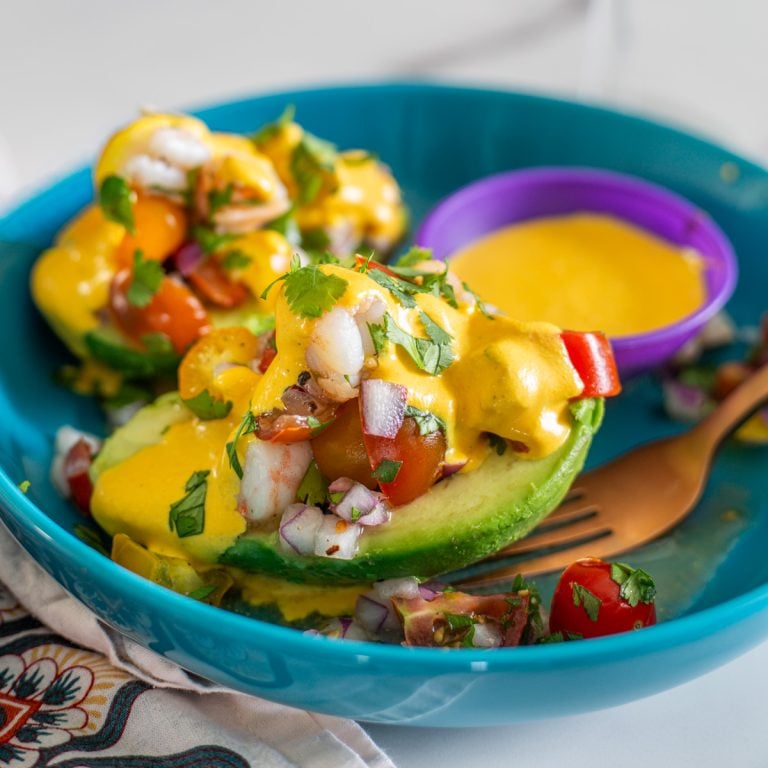 A turquoise ceramic bowl with 2 palta rellena halves, salsa huanca&iacute;na, and copper fork.