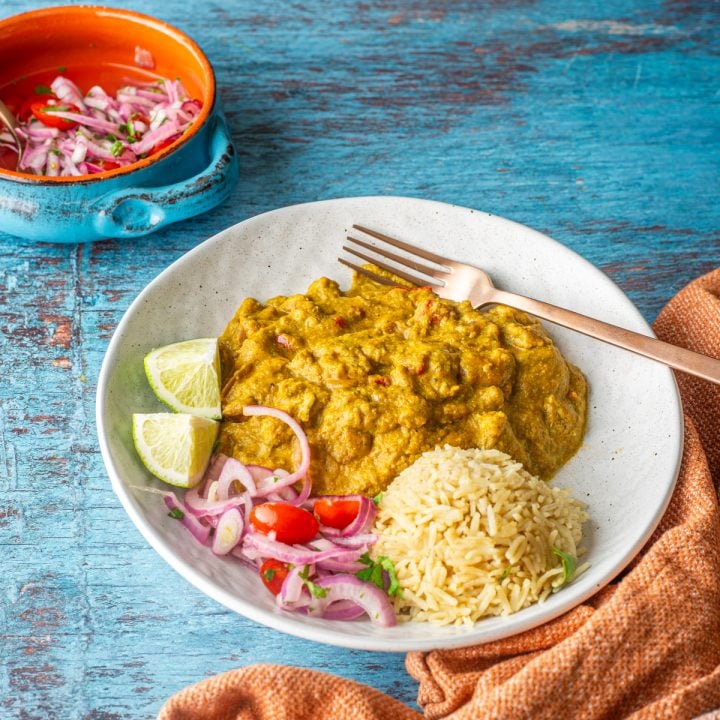 A shallow white bowl with pep&iacute;an de choclo, salsa criolla, and rice with a copper spoon and rust napkin.