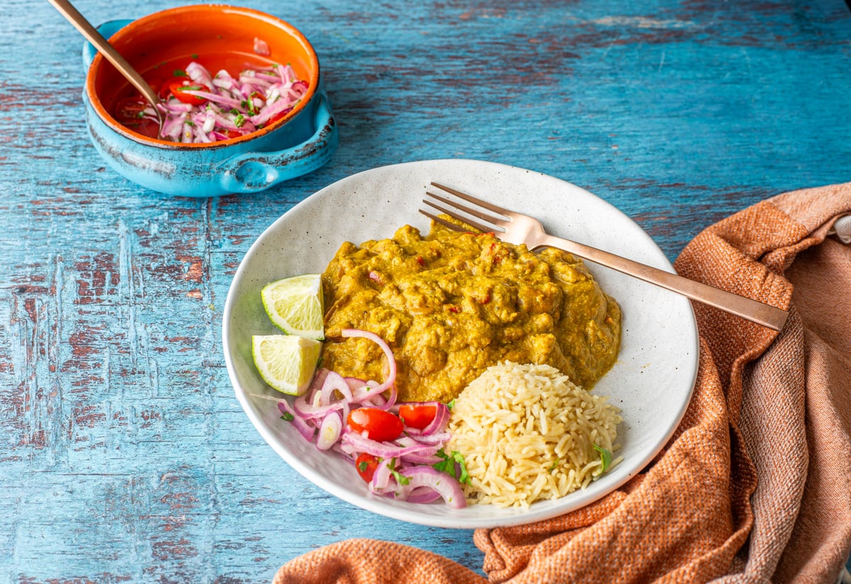 A shallow white bowl with pep&iacute;an de choclo, salsa criolla, and rice with a copper spoon and rust napkin.