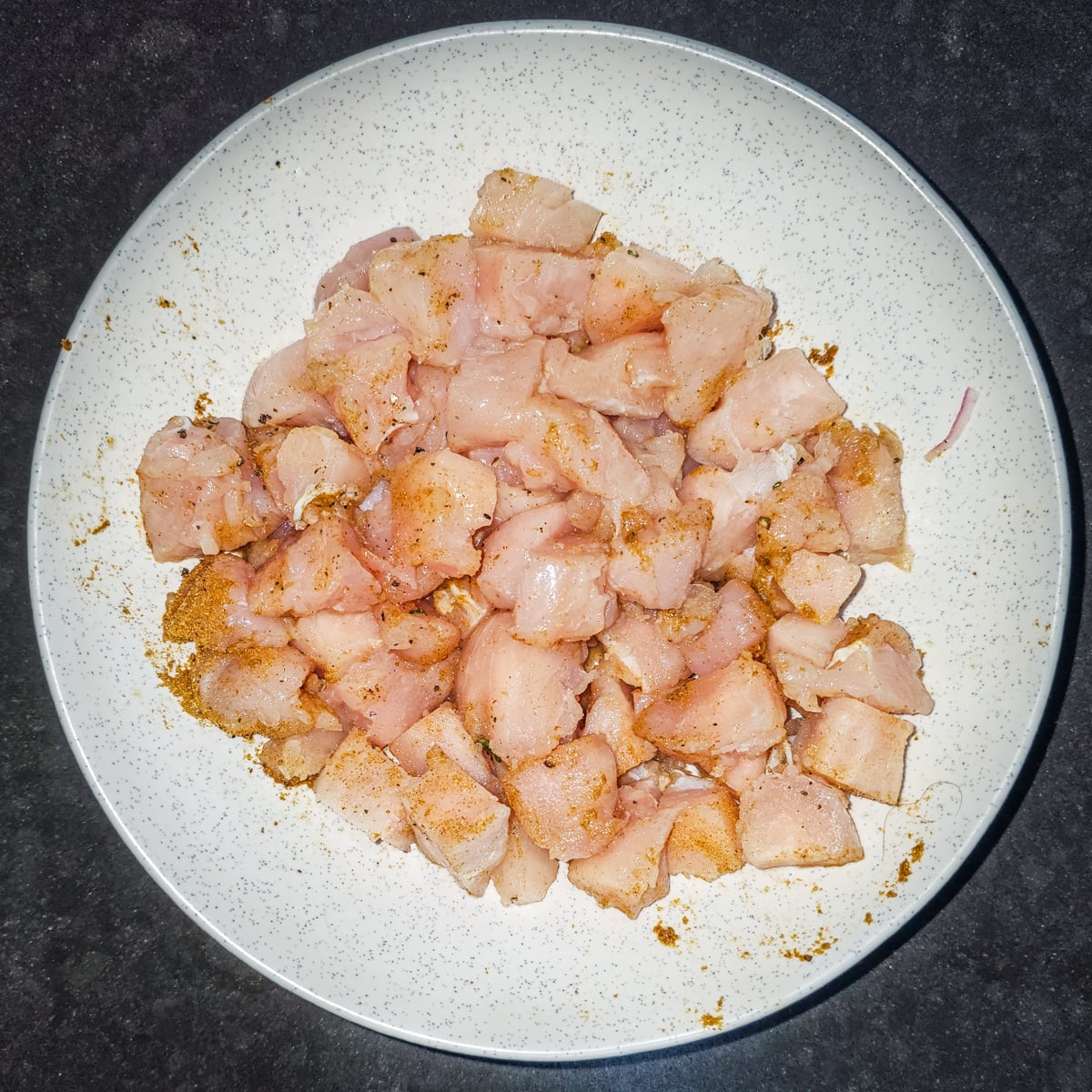 A white bowl with cubed chicken combined with salt, pepper, and ground cumin.