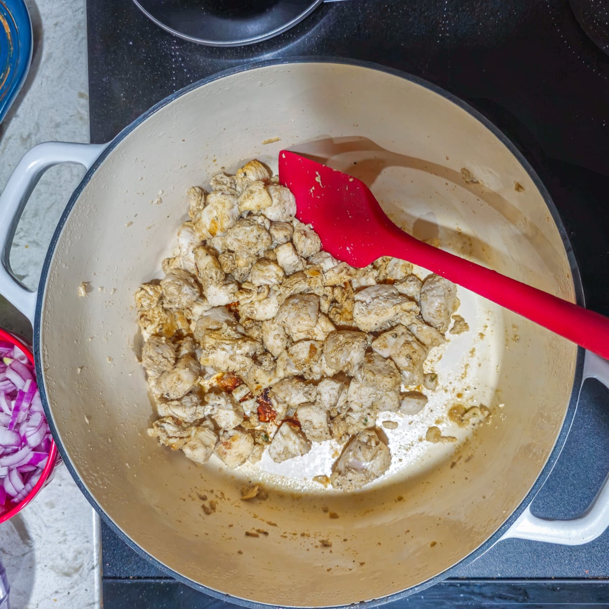 Chicken pieces are browned in a white dutch oven with a red spatula.
