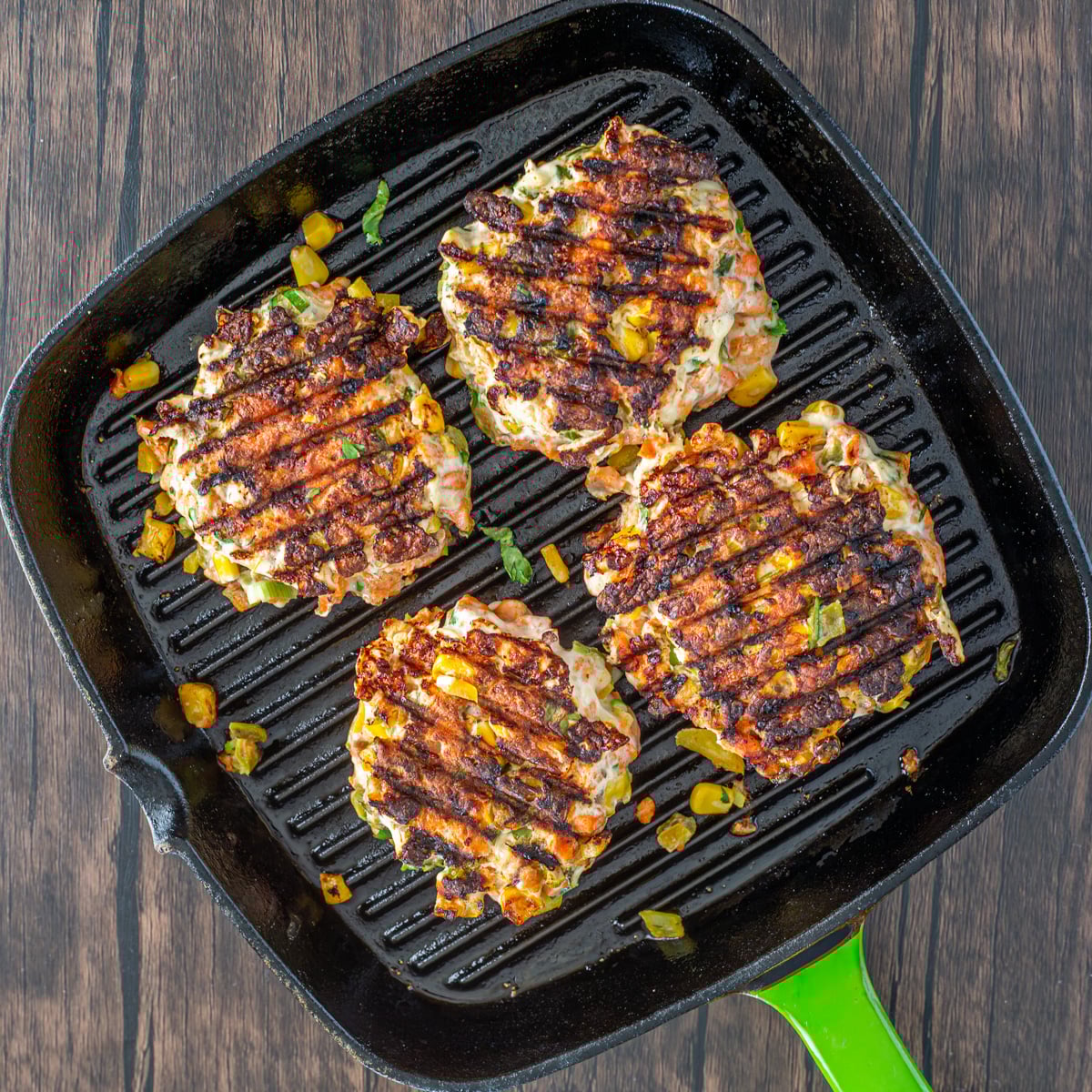 A cast iron griddle pan with a green handle with 4 spicy Mexican salmon patties ready to place on salad.