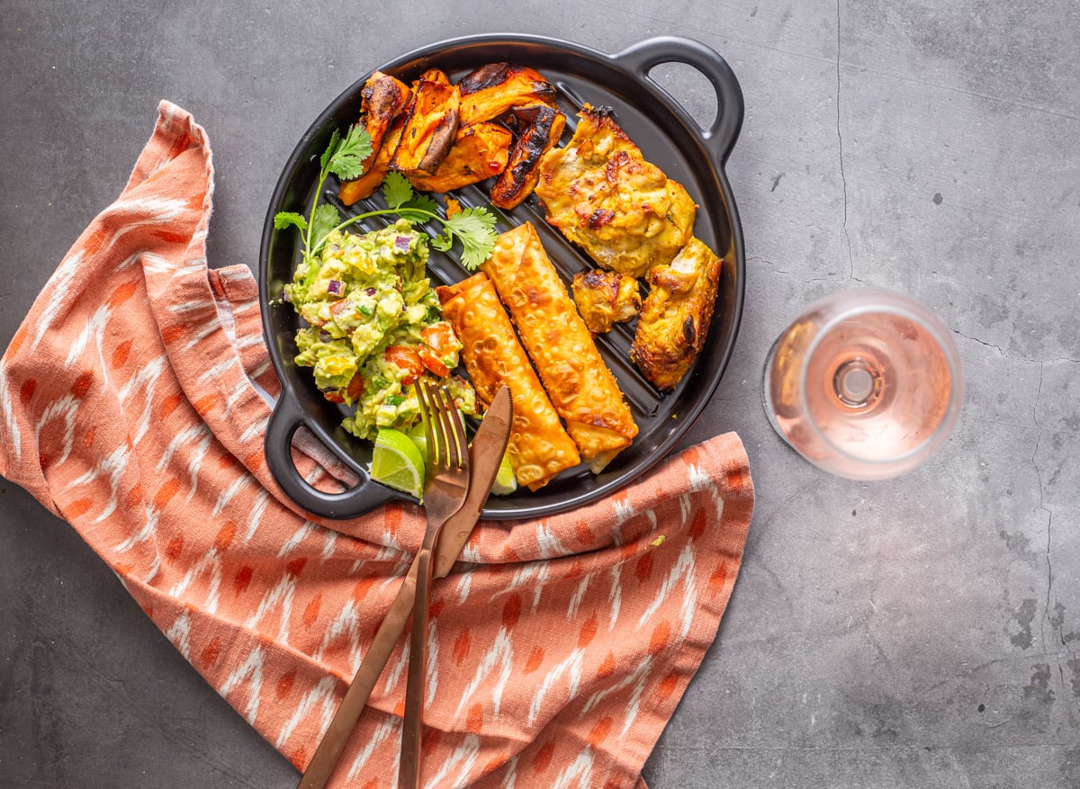 A black ceramic plate with salsa de palta, teque&ntilde;os, and roasted chicken thighs and sweet potatoes.