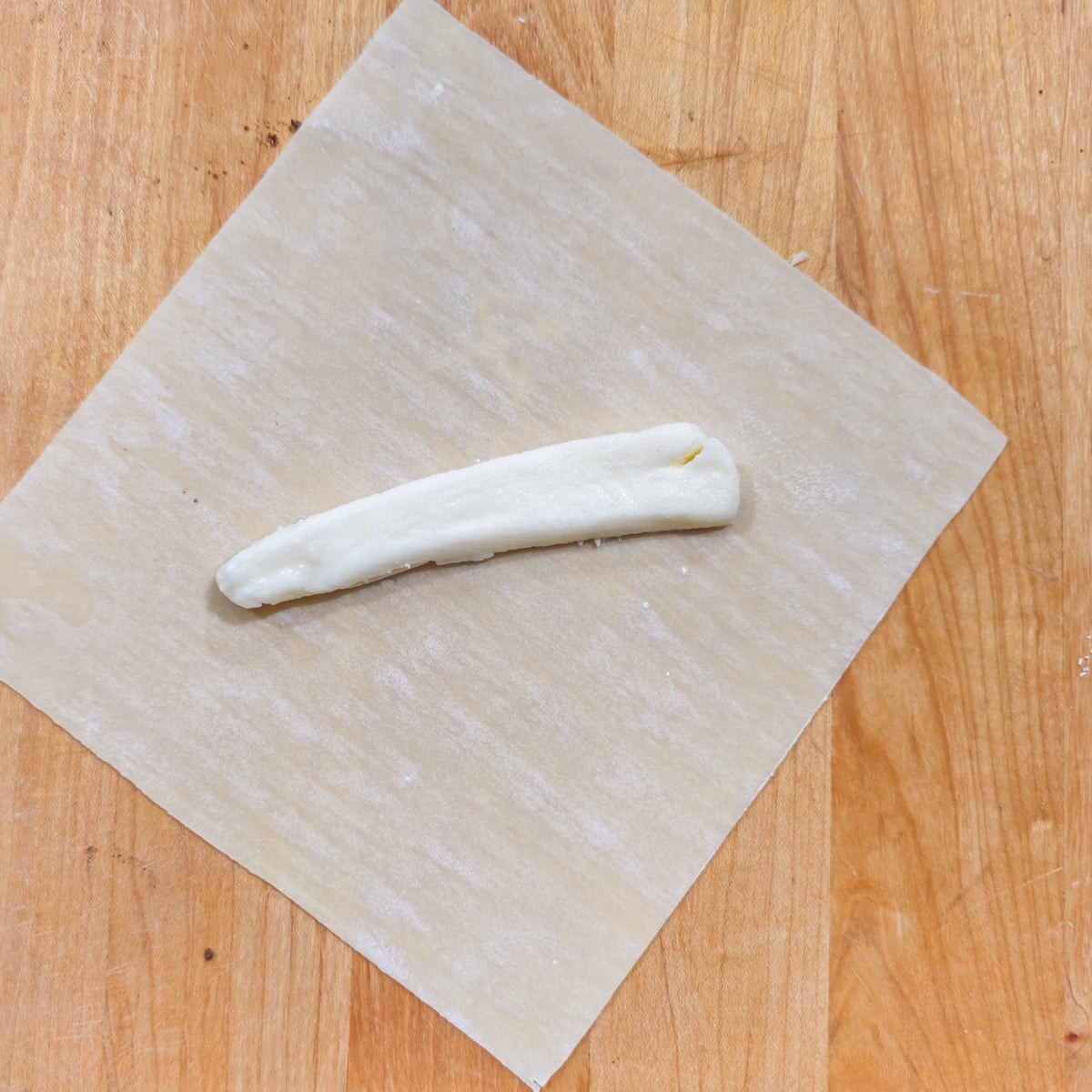 A wood cutting board with a single sheet of eggroll wrapper and a stick of cheese.