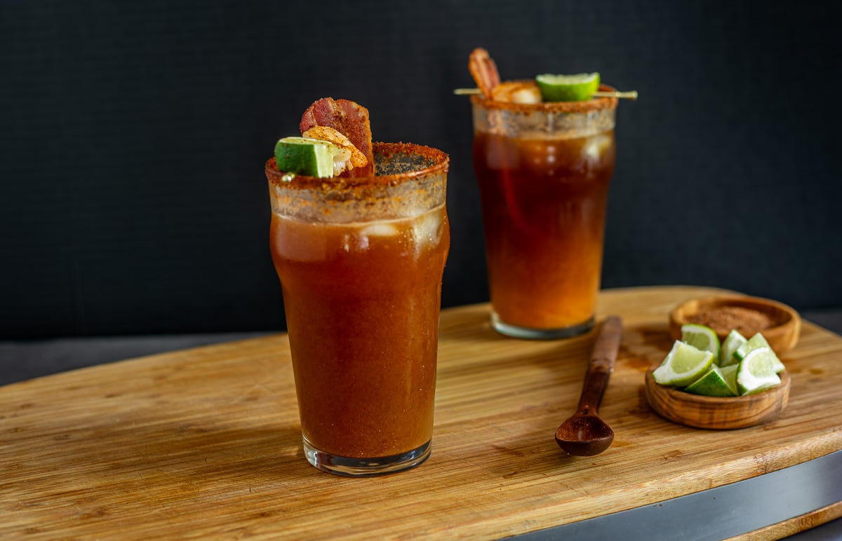 2 garnished miso micheladas on a cutting board with lime wedges.