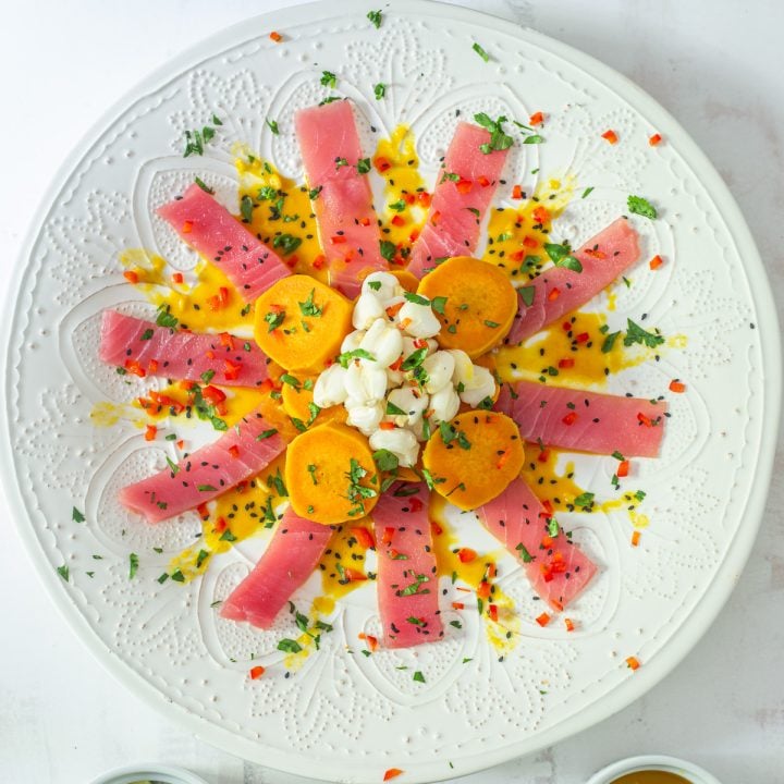 The prepared Tiradito de Atún on an embossed white ceramic plate with sweet potatoes, maiz mote, and garnishes.