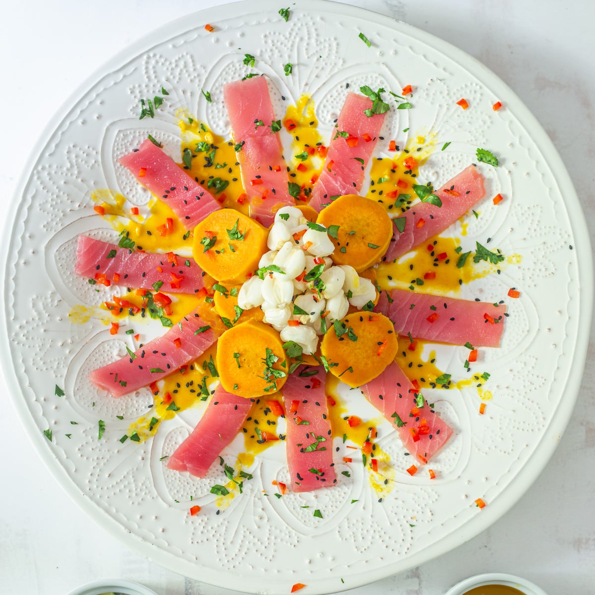 The prepared Tiradito de At&uacute;n on an embossed white ceramic plate with sweet potatoes, maiz mote, and garnishes.