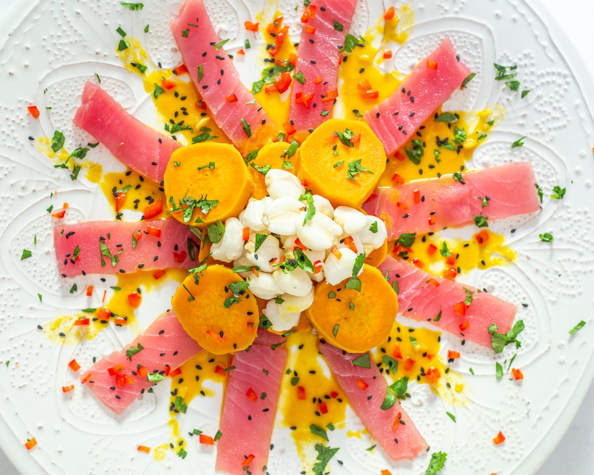 The prepared Tiradito de At&uacute;n on an embossed white ceramic plate with sweet potatoes, maiz mote, and garnishes.