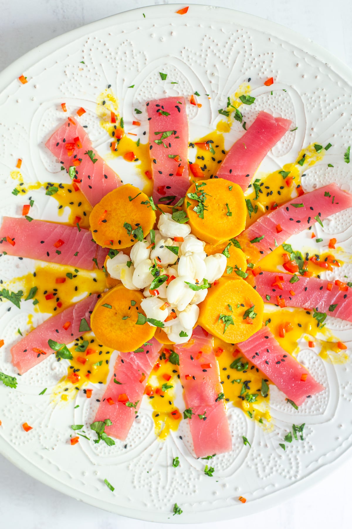 A white plate of Tiradito de At&uacute;n with sweet potatoes, maize mote, aj&iacute; amarillo sauce, herbs, and black sesame seeds.