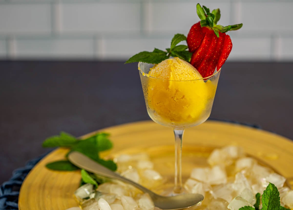 A Nick and Nora glass with a creamy scoop of passion fruit sorbe on a yellow plate with ice cubes and mint.