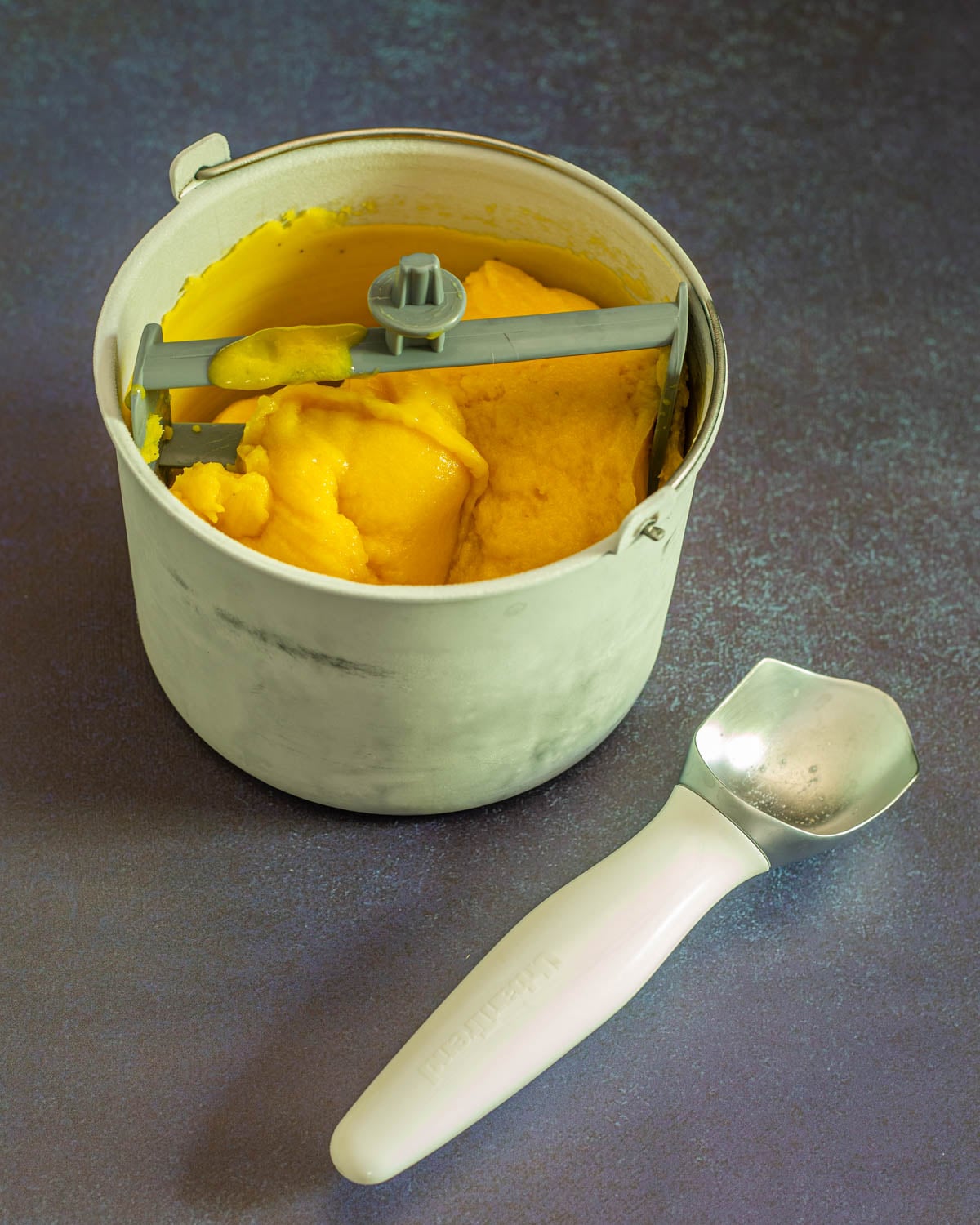 The freezer bowl of beautiful passionfruit sorbet with mango alongside a white ice cream scoop.