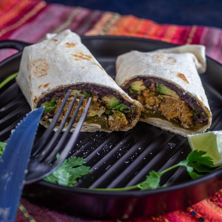 A black ceramic plate with 2 meatloaf burrito halves with a fork and colorful napkin.