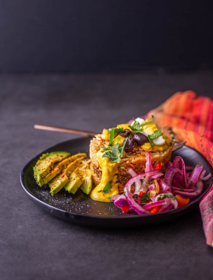 A black stoneware plate with a single serving of arroz tapado with salsa criolla and sliced avocado.